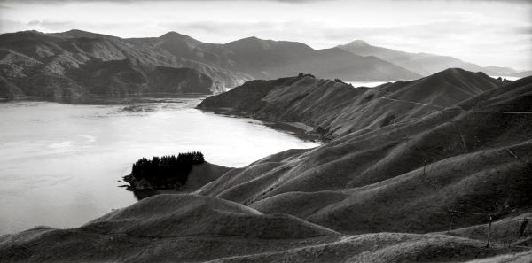 Black and white photograph taken from the top of a curving hillside, overlooking a tranquil sound, flanked by angular forested hills.