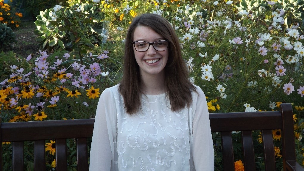 A portrait of a young woman with shoulder length brown hair, wearing glasses, sitting on a bench in a park