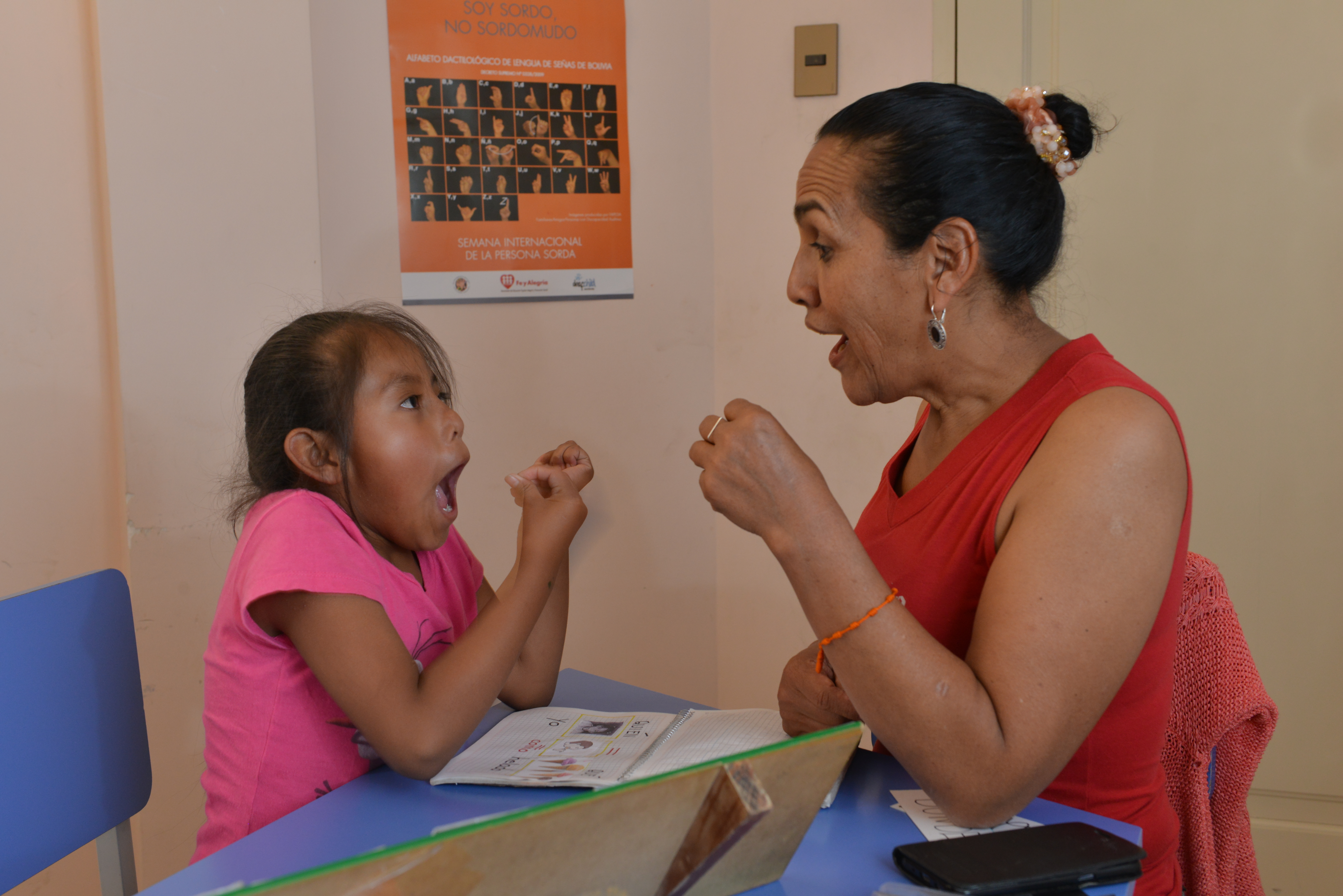 A girl is sitting opposite a woman. The girl is gesticulating as she learns sign language. She is gesturing with her hand and making a shape with her mouth.