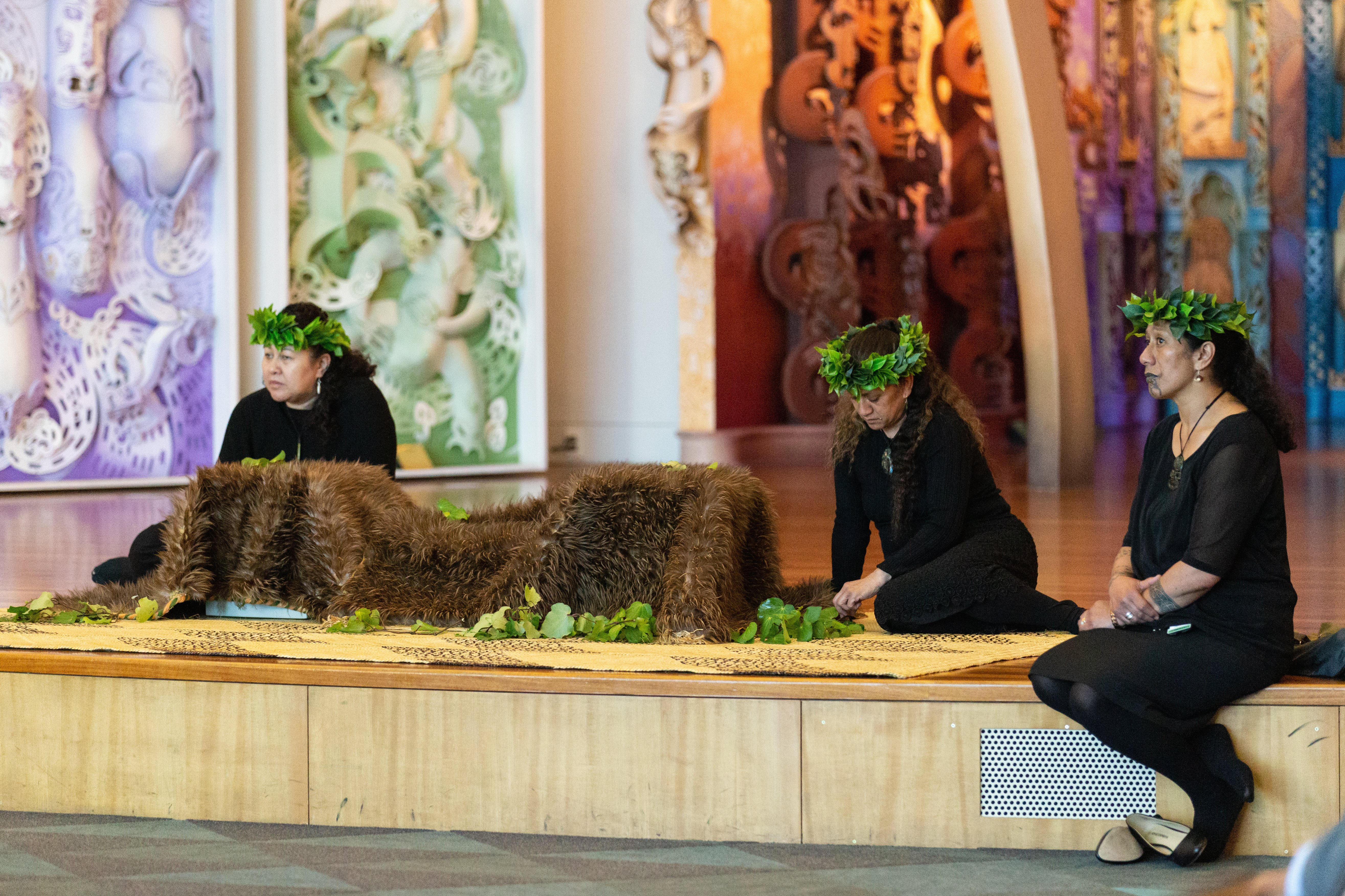 Three Māori women are seated in a carved marae (meeting house). They wear black, with green woven wreaths on their heads. Some of them have traditional facial tattoos. Before them a feathered cloak is draped over boxes containing repatriated remains,