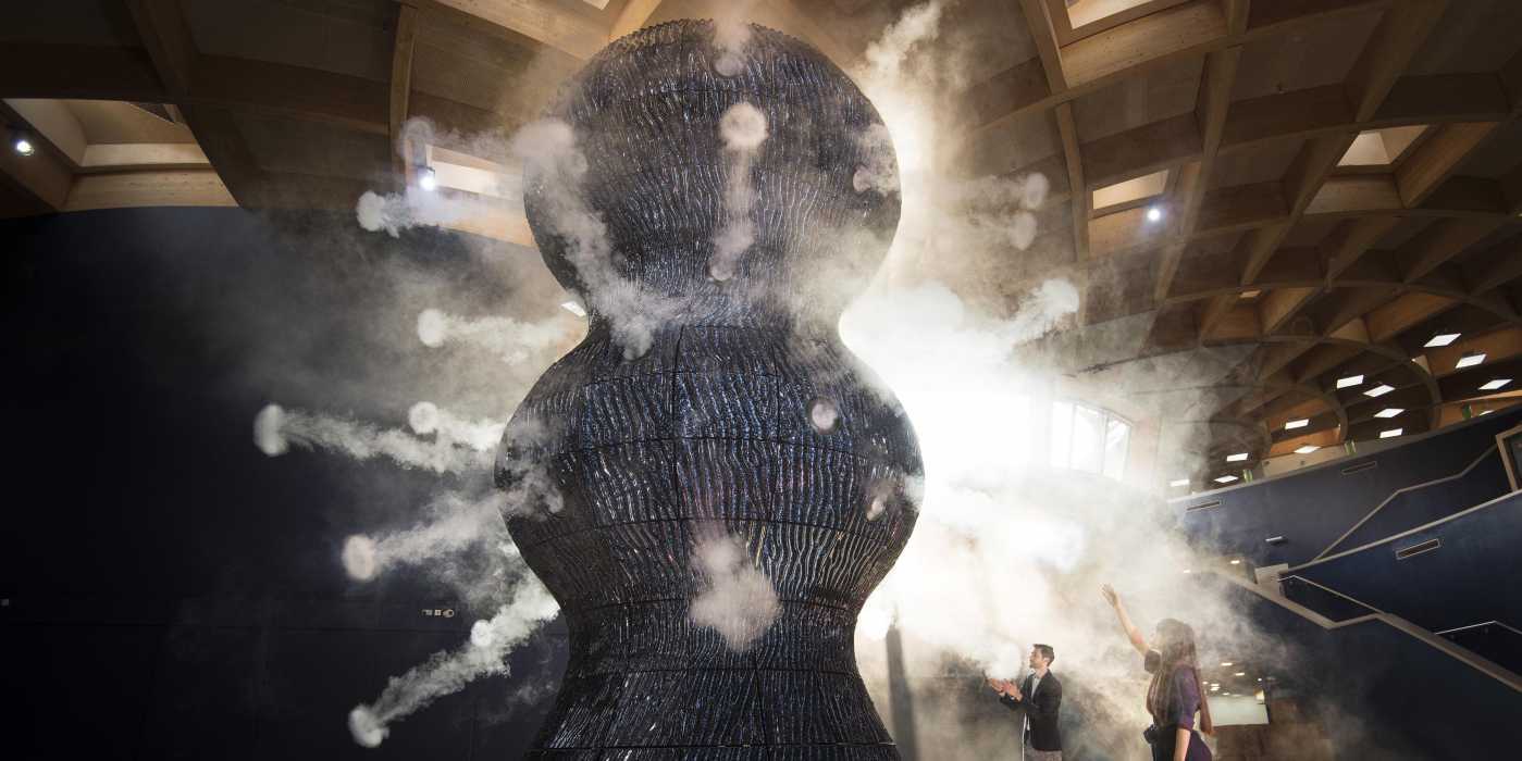 Infinity Blue Sculpture. An 8.5-metre high sculpture representing cyanobacteria. From the sculpture, vapour rings are cast in all directions, representing the oxygen that cyanobacteria release. Two people are holding their hands up to the oxygen rings.