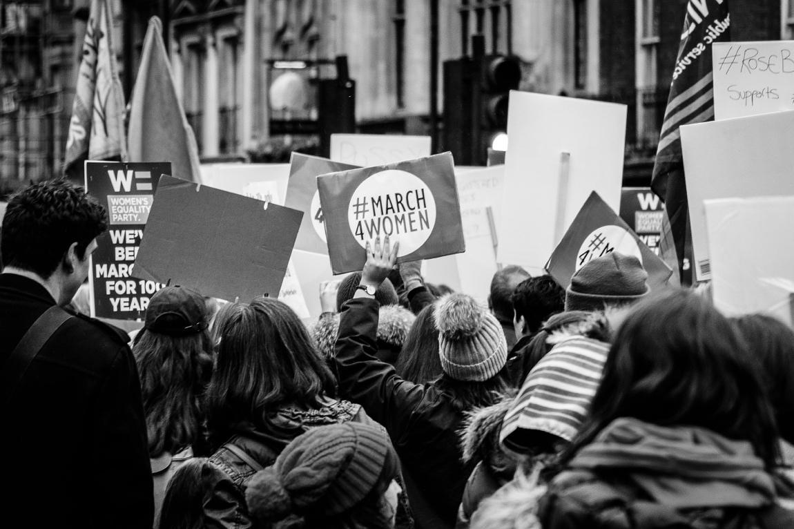Black and White photo of a march, taken from behind. Banners say #MARCH4WOMEN