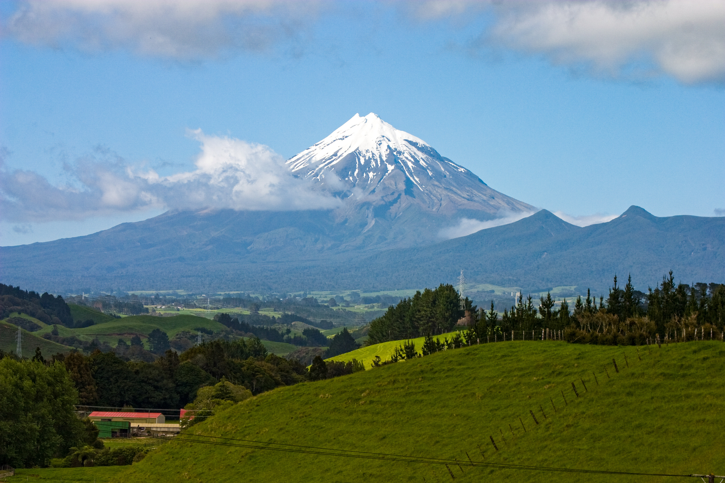 Landcape picture of Mount Taranaki, a snow-capped volcano in rolling green farmland. the mountain is surrounded by a ring of Native forest, surrounded by farmland.