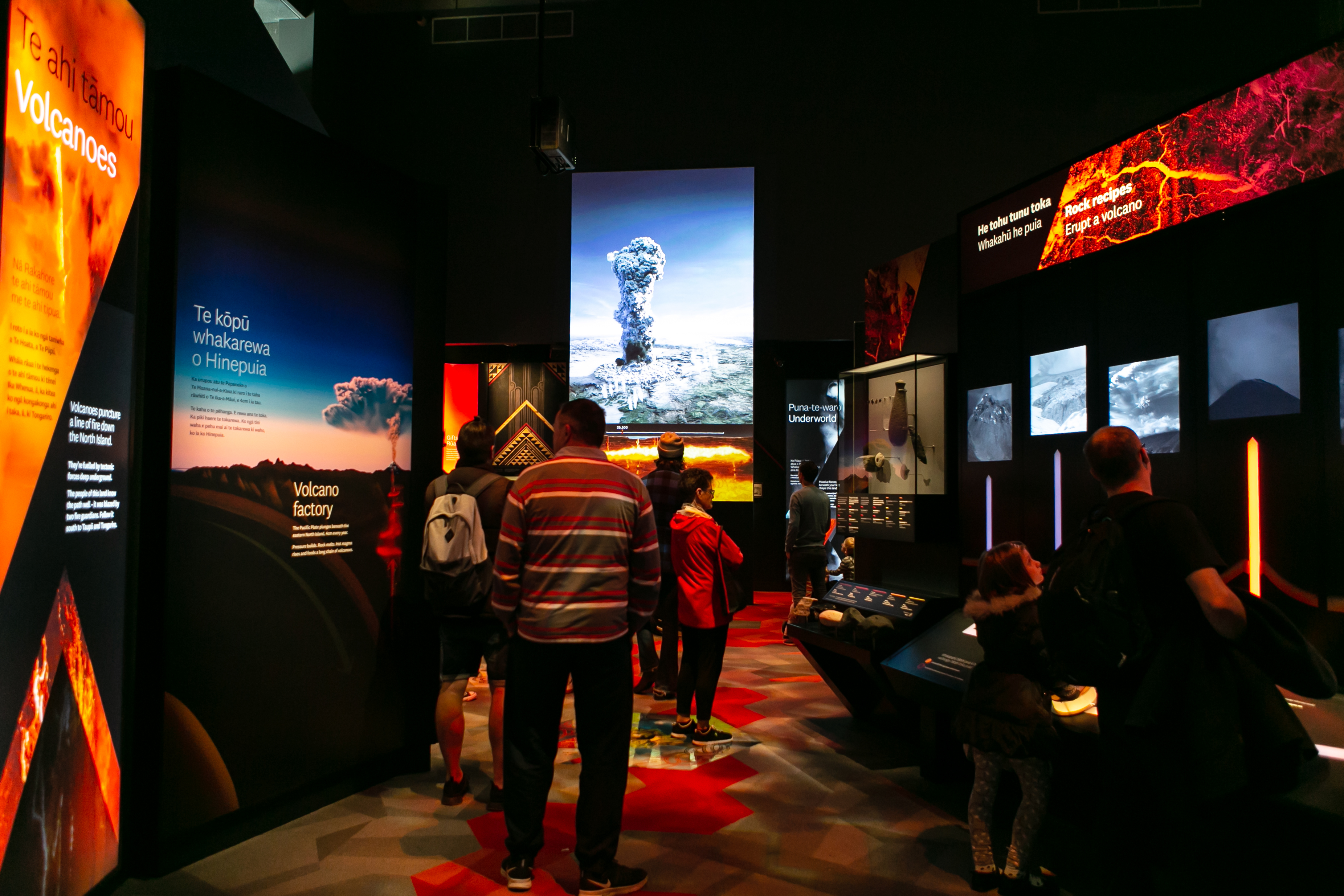 a family walks through an exhibition on volcanoes. On the left is information panels and text, ahead is a large image of an ash plume, and to the right is a man and child pressing buttons on an interactive of how lava flows in volcanoes