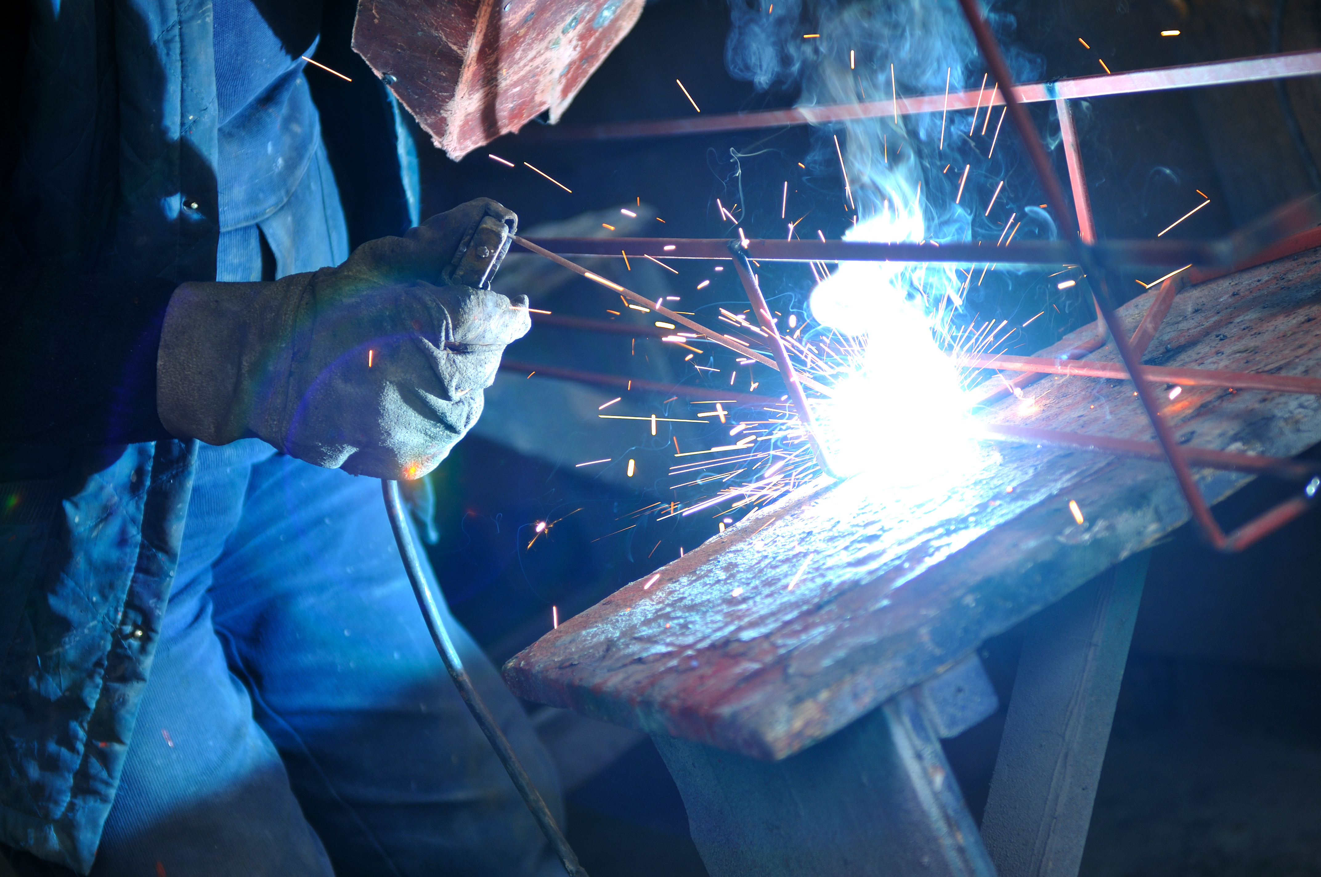 The hands of a welder is shown, with welding light and fumes present.
