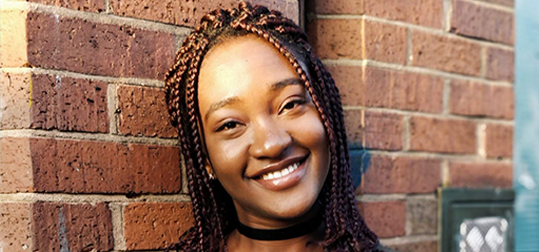 A portrait of a young woman with long plaited hair standing in front of a brick wall