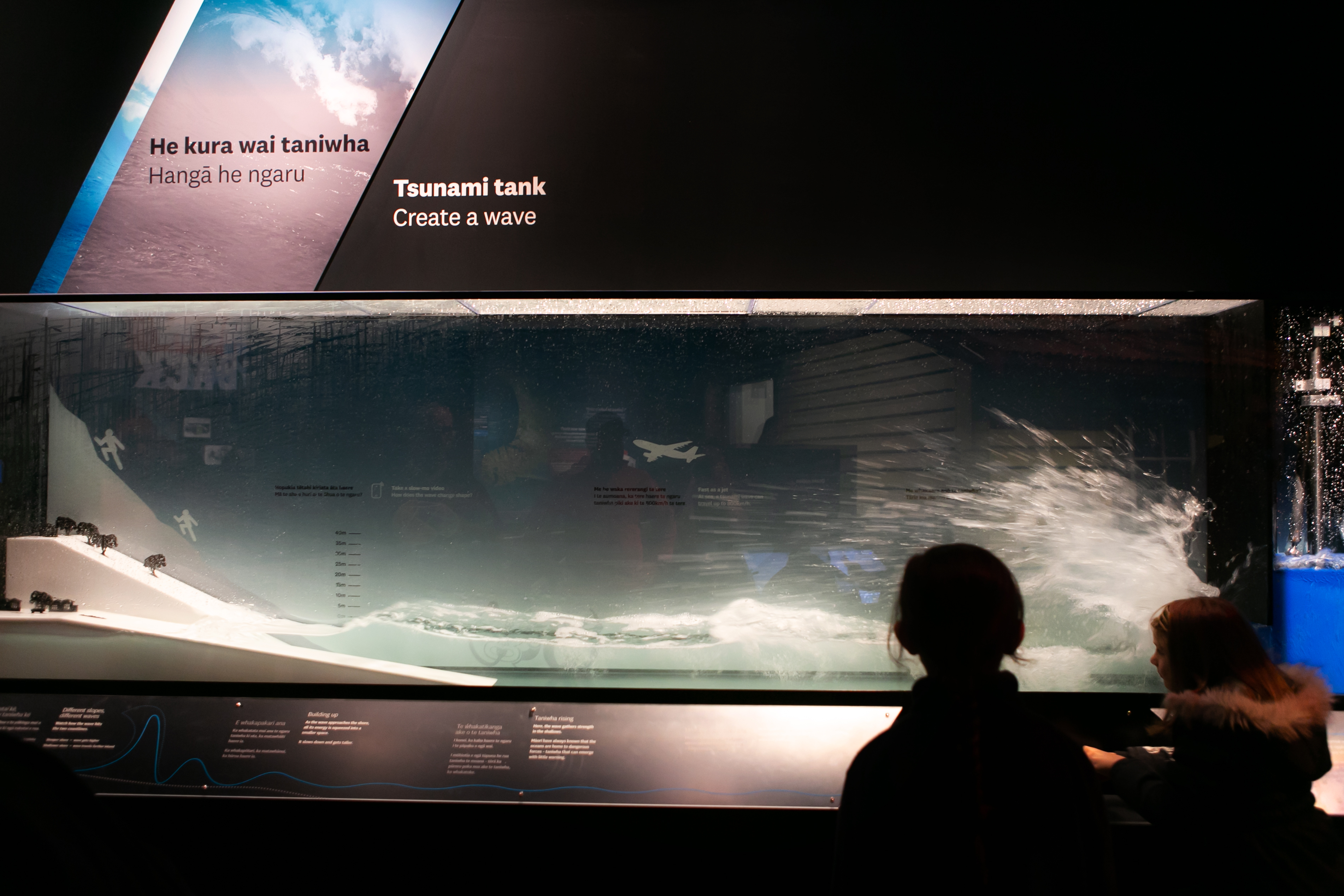 a child stands silhouetted in front of a long illuminated tank of water in which they have set off a miniature tsunami over a diorama of the coastline