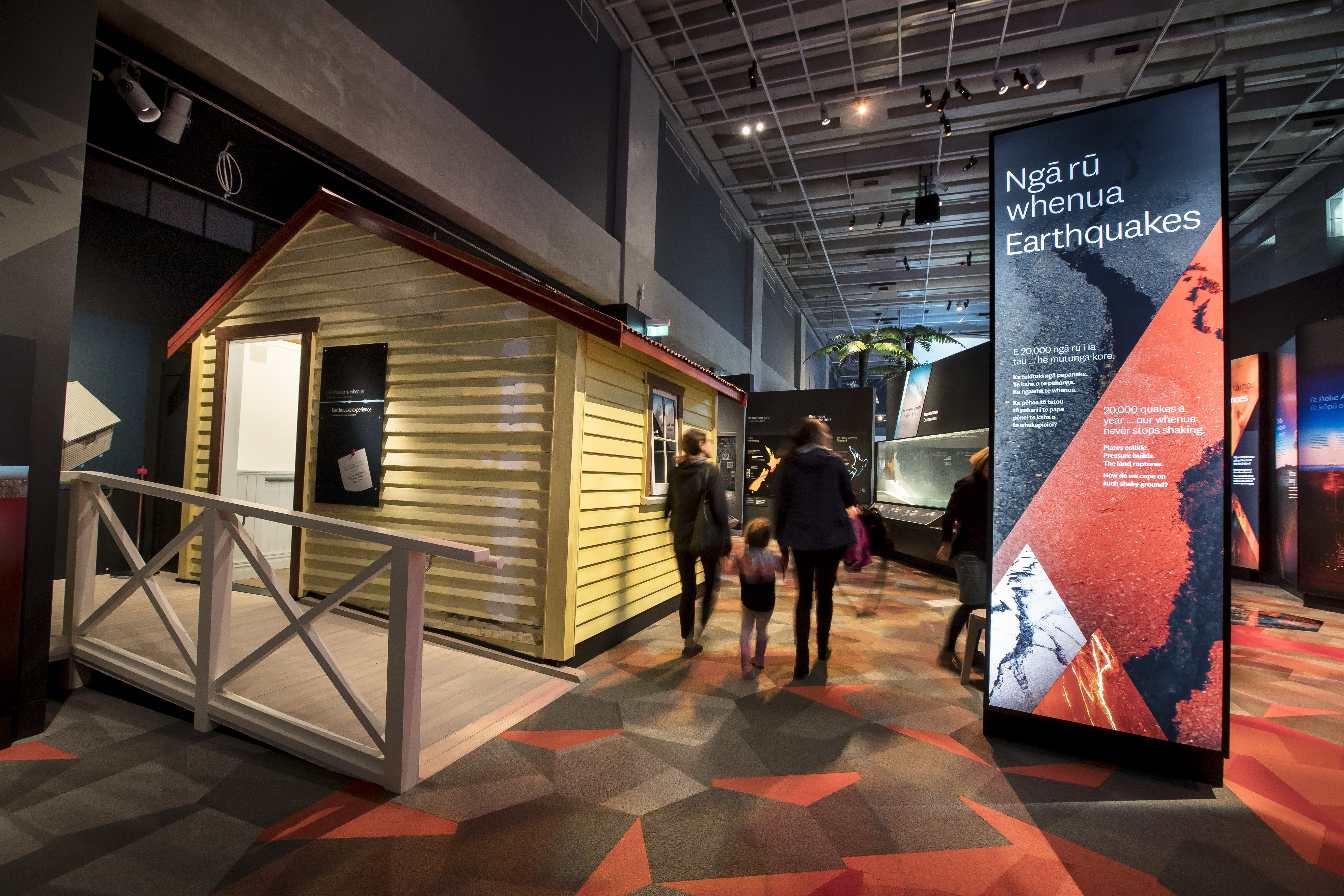 A family walks through the Active Land exhibition, an informative banner about earthquakes is to their right, and to their left is a small weatherboard shack painted yellow- the 'Quake House'.