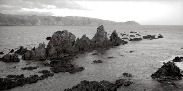 Black and white photograph looking out into a harbour and the distant sea. A peninsula juts out in the distant background, while the foreground features sharp rocks jutting high out of the ocean in a line, like a row of sharp teeth.