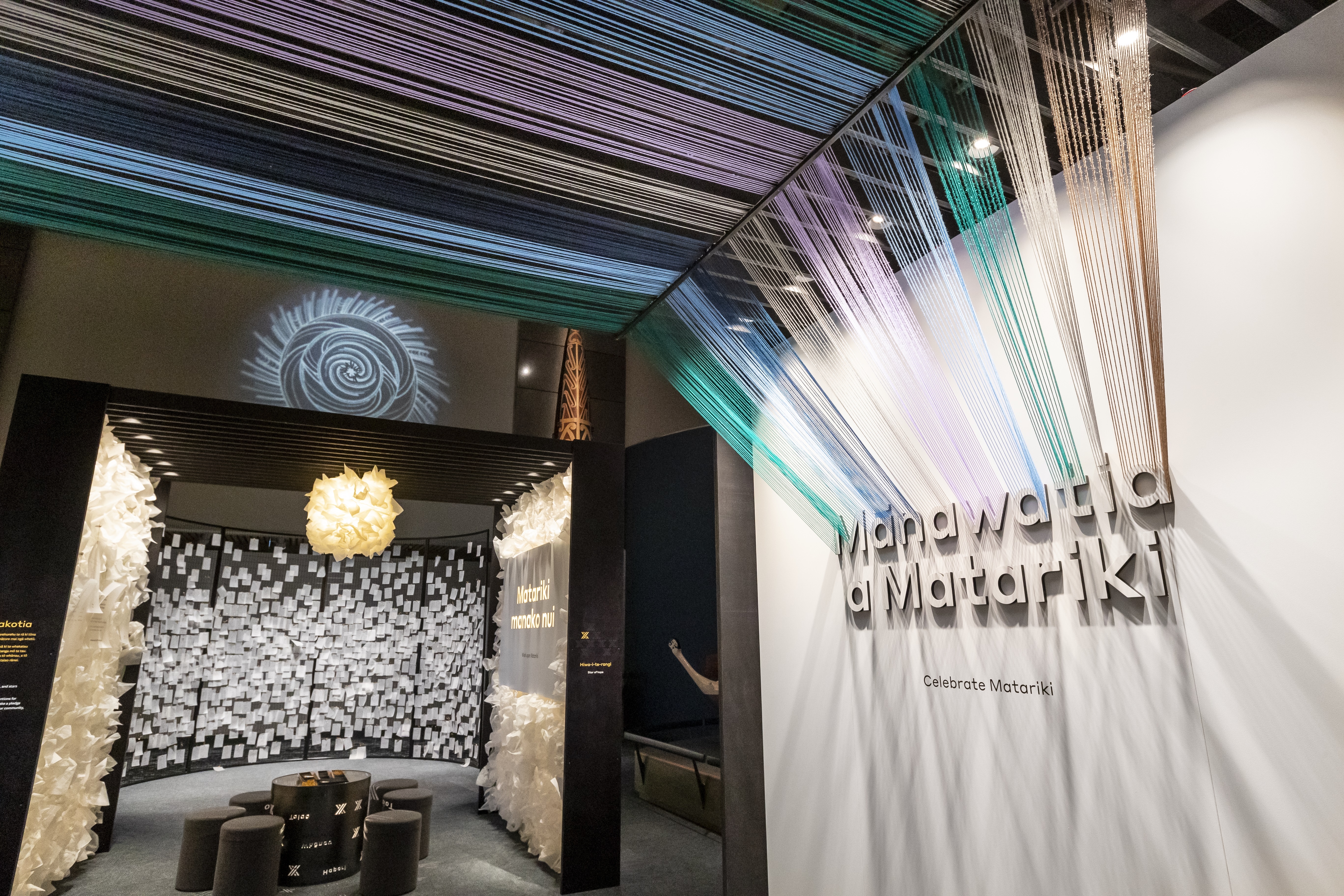 The Matariki exhibition at Te Papa. A wall says Mānawatia a Matariki in floating white letters, hundreds of strands ov coloured string arc over like a rainbow. Beyond is a small table and chairs and a wall full of white scraps of paper- pledges for the new year from visitors.