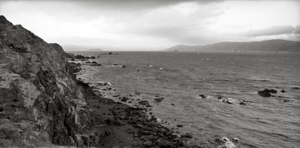 Black and white photograph looking from a rocky shore towards a far peninsula. Two small islands can be seen through a distant mist.