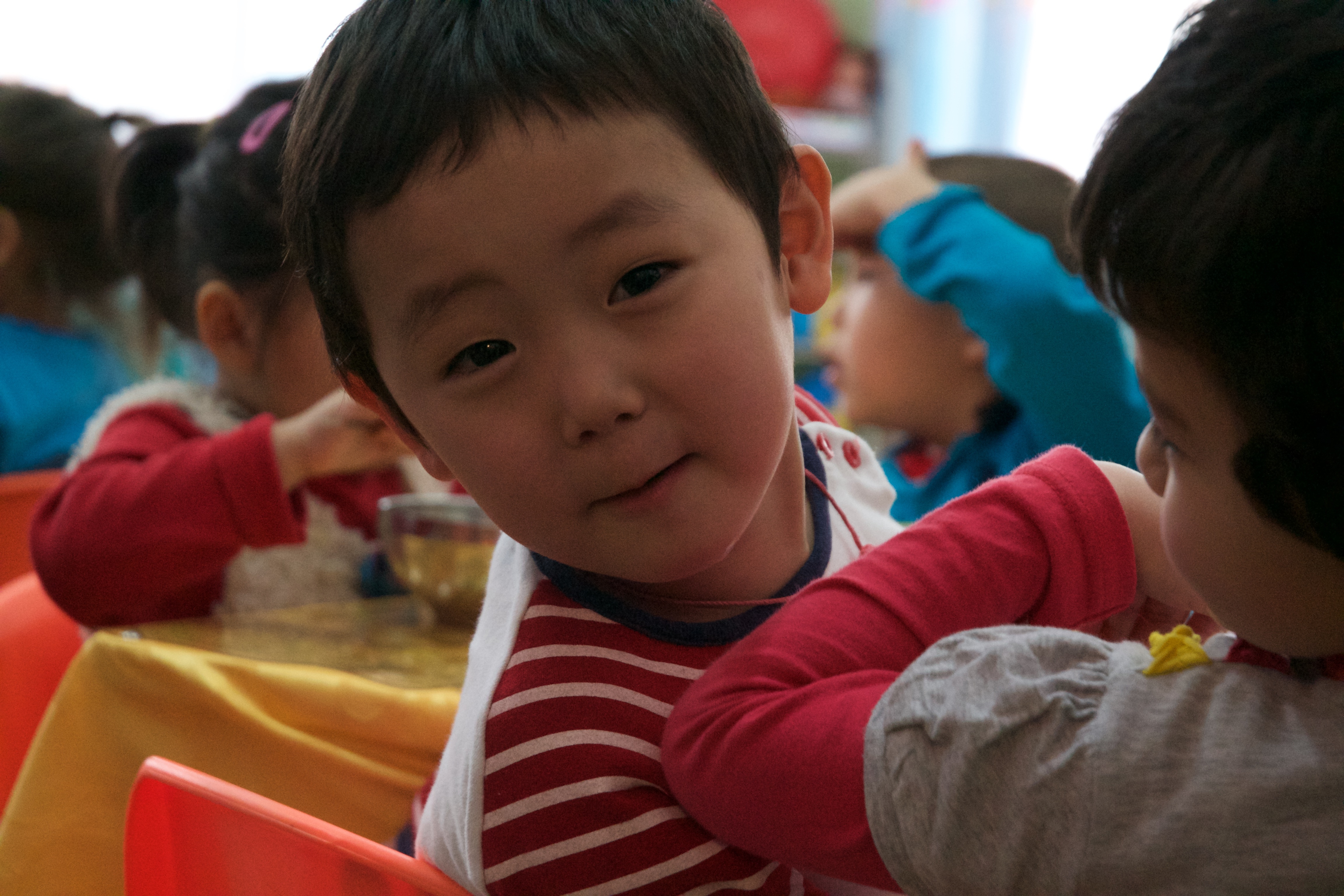 A boy leaning back to smile at the camera as he sits at a table with other children.