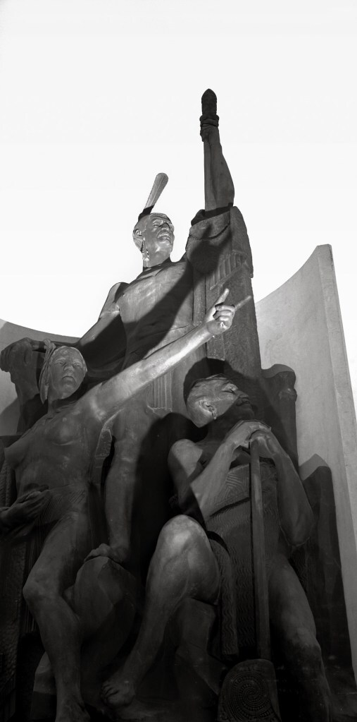 Black and white photograph of a statue featuring three Māori figures, taken from below. A chiefly looking man stands highest, a decorative pin in his topknot. stands highest, gazing outwards and grasping a staff. A bare-chested woman stands below him, pointing into the distance. Below her, another man in traditional dress looks towards where she points, resting his hands on the head of his staff.