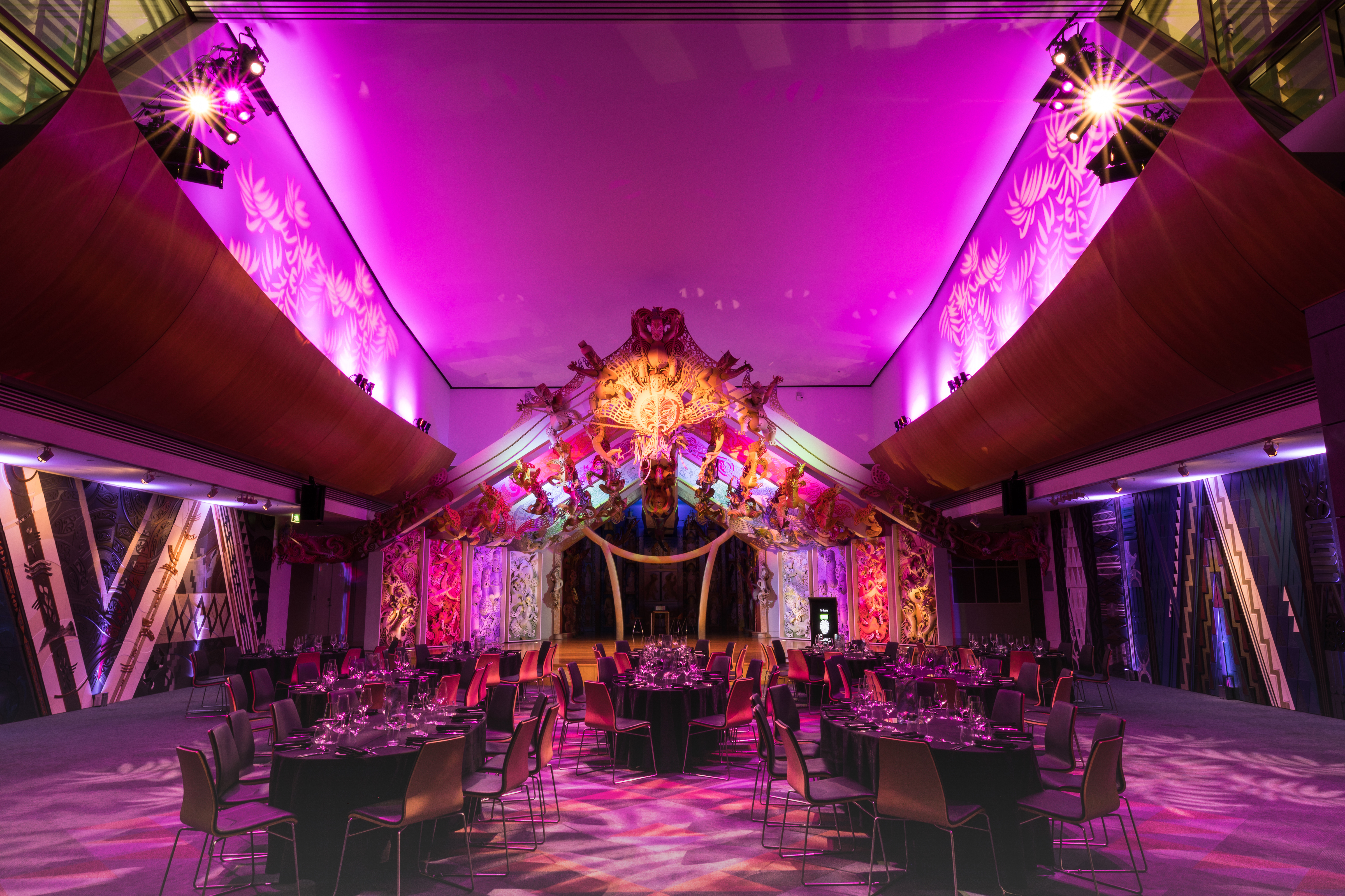 A wide view of the marae, set for a formal event. bright pink light glows above the space, with the wharenui at its centre and formal dining tables set up in the ātea