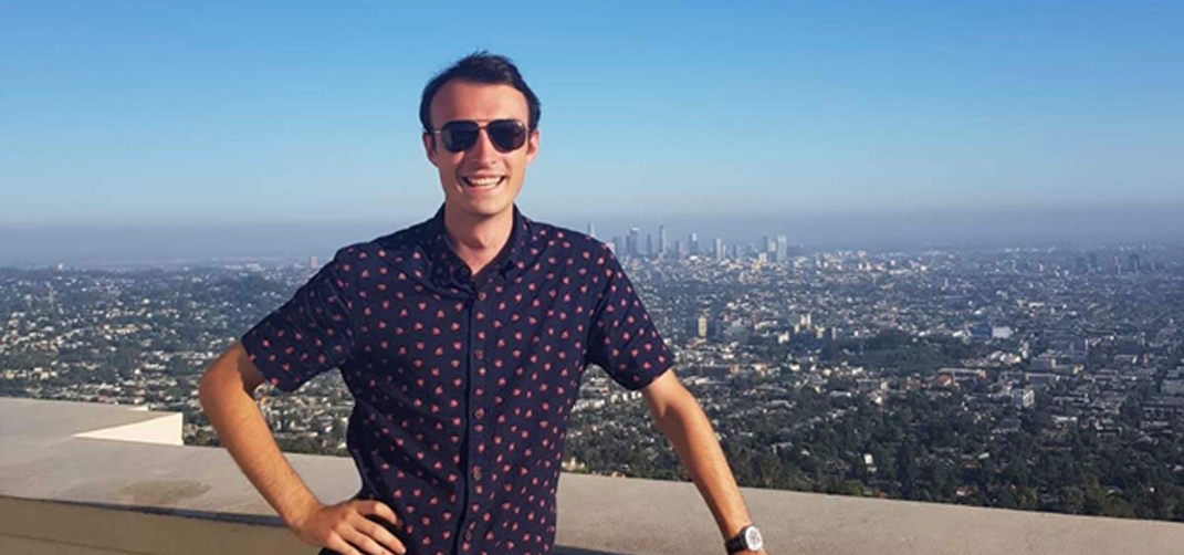 A young man wearing sunglasses is smiling as he stands with one hand on his hip and the other on a balcony overlooking a cityscape