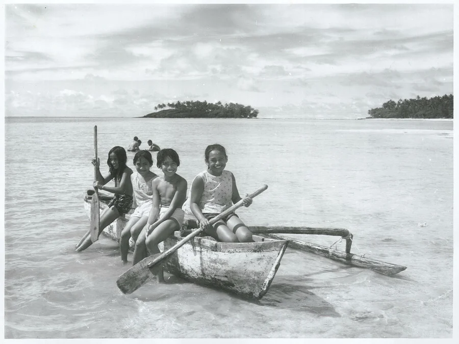 A black and white photo of four pacific island children seated on a small outrigger canoe in shallow water with an island in the background