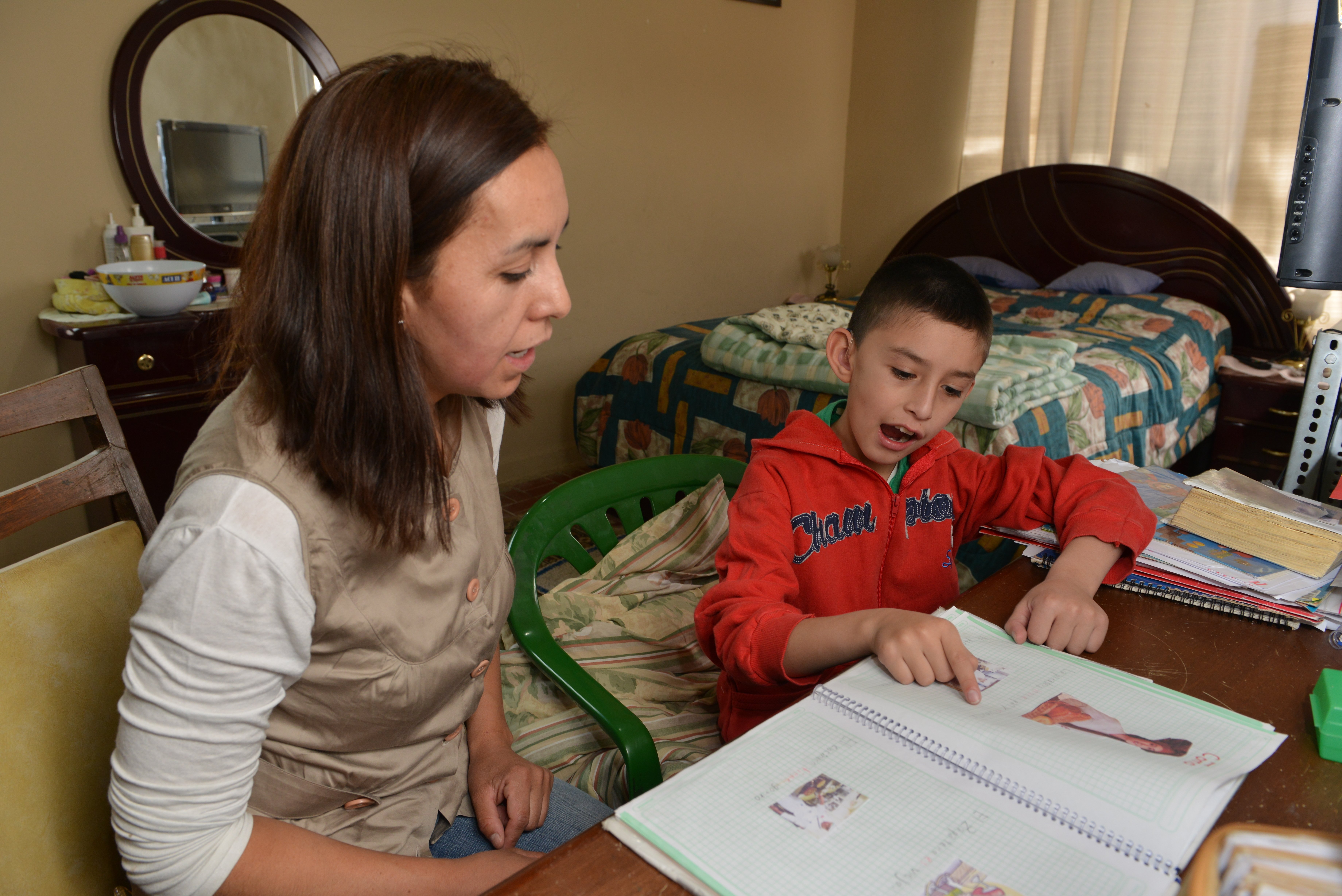 A boy with a hearing impairment is pointing at a book with images in. He is being supported by a woman. They are in a bedroom