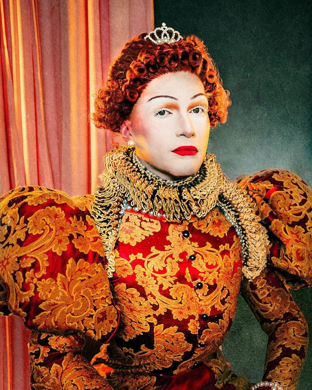 Mary has bright red, tightly curled hair and a small crown with diamonds and pearls. Her face is pale with bright red lipstick and orange and green eye shadow, and she faces out at the viewer. Her dress is elaborate, with red silk and decorative orange flowers. The sleeves of the dress are puffed up at the shoulders. The is a ruffled lace collar in gold around the neckline and extending down.