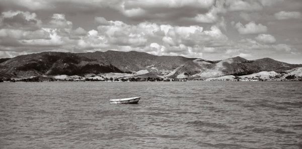 Black and white photograph of a small rowboat floating in a broad harbour, with rolling hills in the background and fluffy clouds filling the sky.