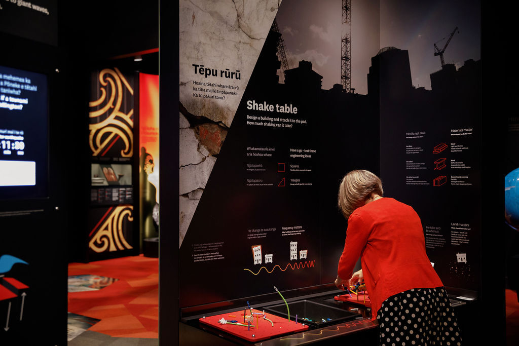 a woman in red stands before an interactive exhibit. walls of pictures and text surround her. She uses bright plastic connectable pieces to build a structure on top of a small red surface that will simulate a small scale earthquake when a button is pushed