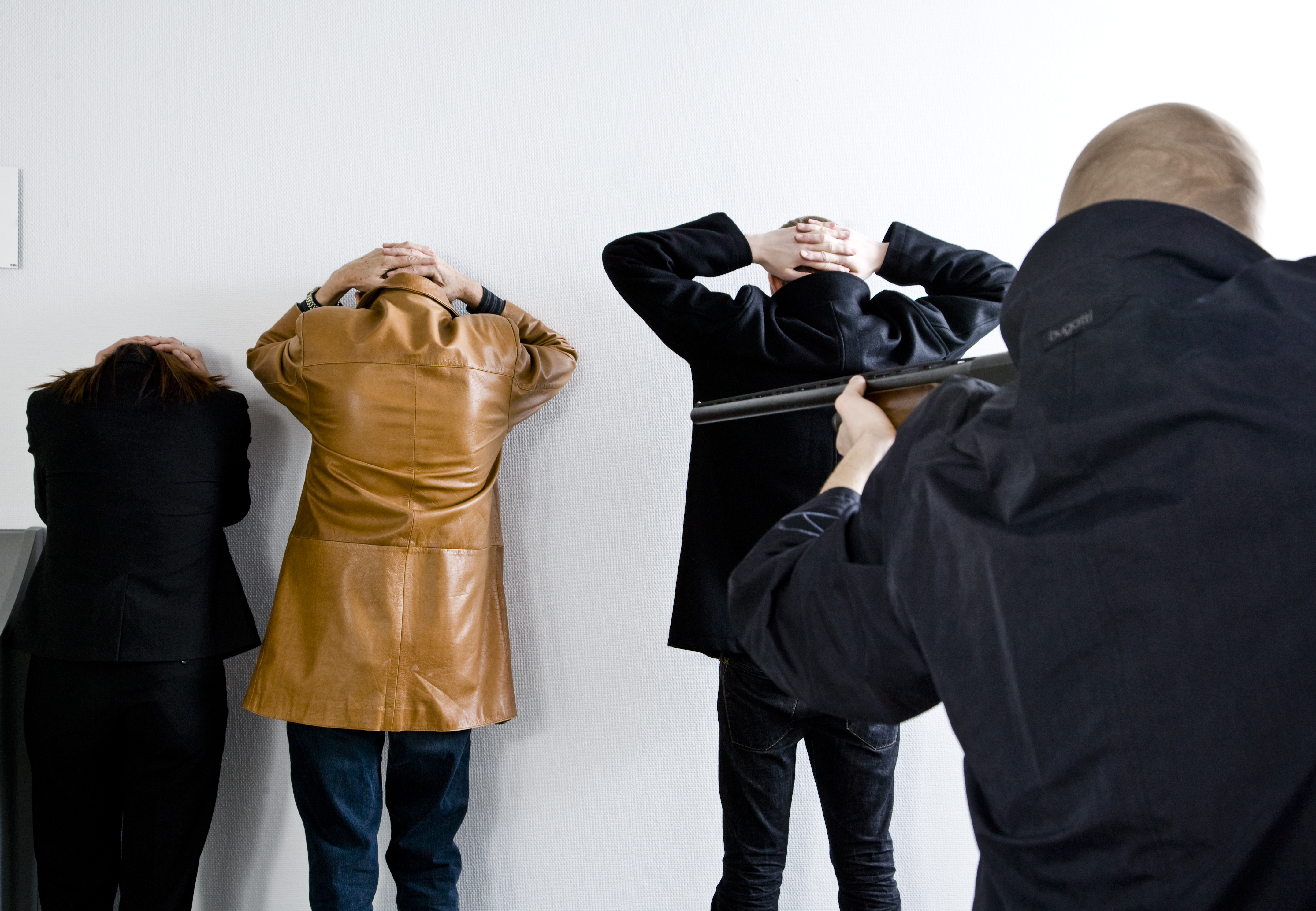 Three people standing againt the wall with their hands behind their head, while another is pointing at them with a gun