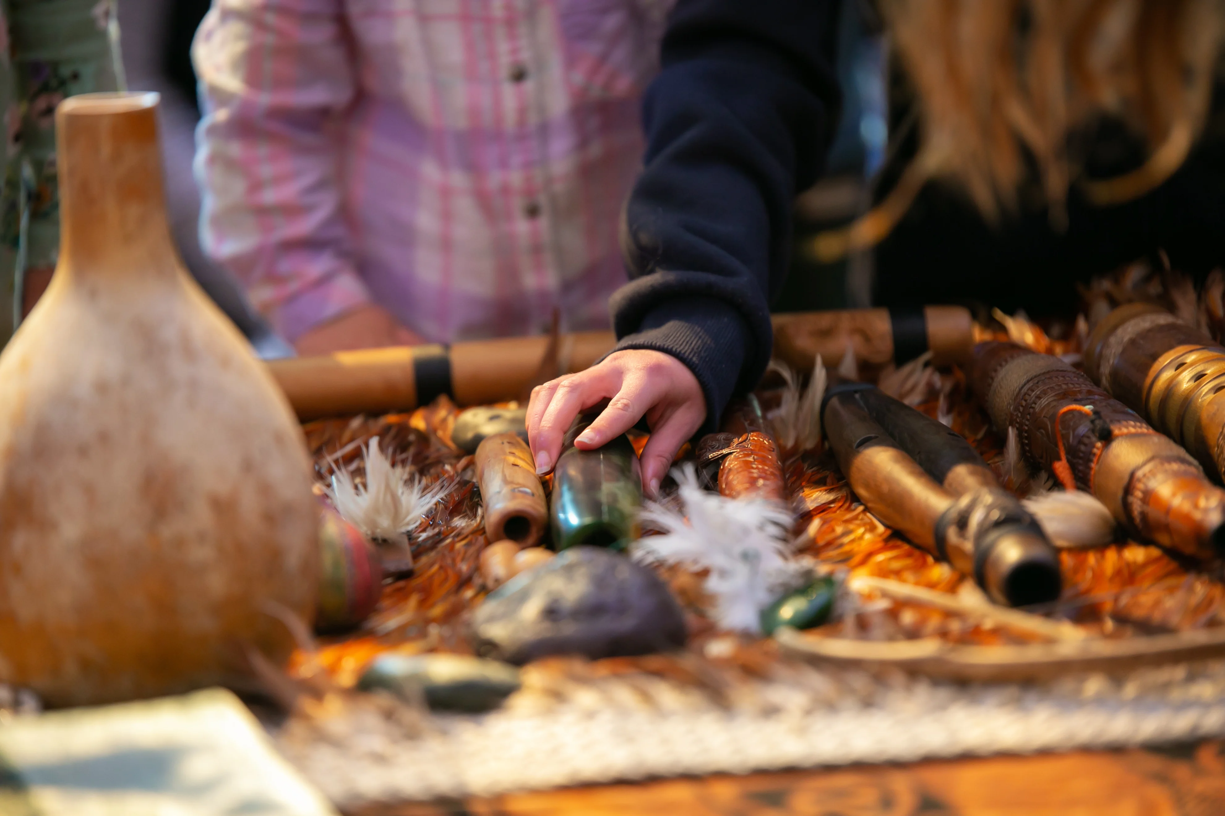 A childs hand rests on an item on a table of pūoro (Māori musical instruments) made with carved wood, stone, and feathers.