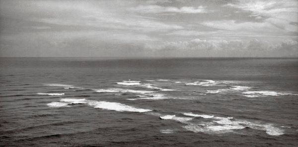 Black and white photograph over a vast ocean. In the foreground we see white caps of swirling waters. the waves on each side of the white caps are running in different directions, marking the collision of two seas.