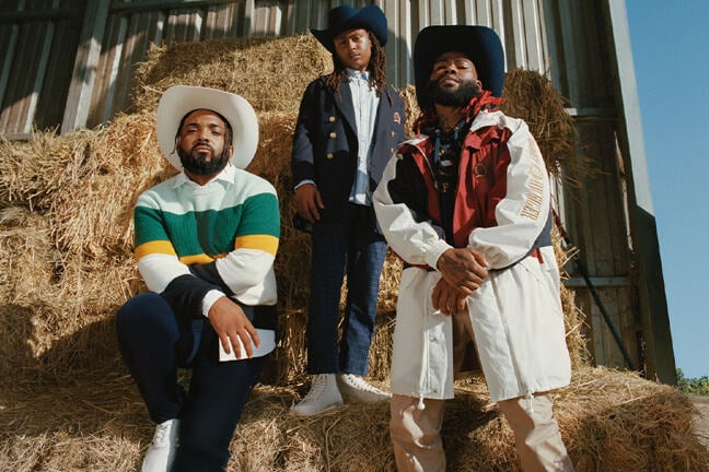 A photograph of three members of the Compton Cowboys against a backdrop of hay bales in a warehouse. The Cowboy on the left is leaning against the hay bales with a knee raised, the Cowboy in the middle is standing on top of a hay bale whilst the Cowboy on the right is standing upright. All three are looking in to the camera