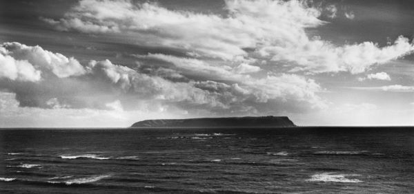 Black and white photograph of a long, flattened island in the distance. Huge banks of cloud roll along the sky.