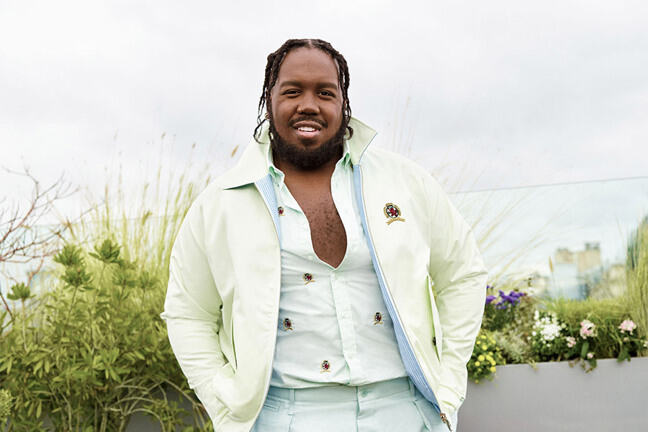 A photograph of Kiddy Smile standing in front of a row of plants and flowers with a city in the background. Kiddy is wearing a cream coloured jacket with a white shirt underneath and pale blue trousers.