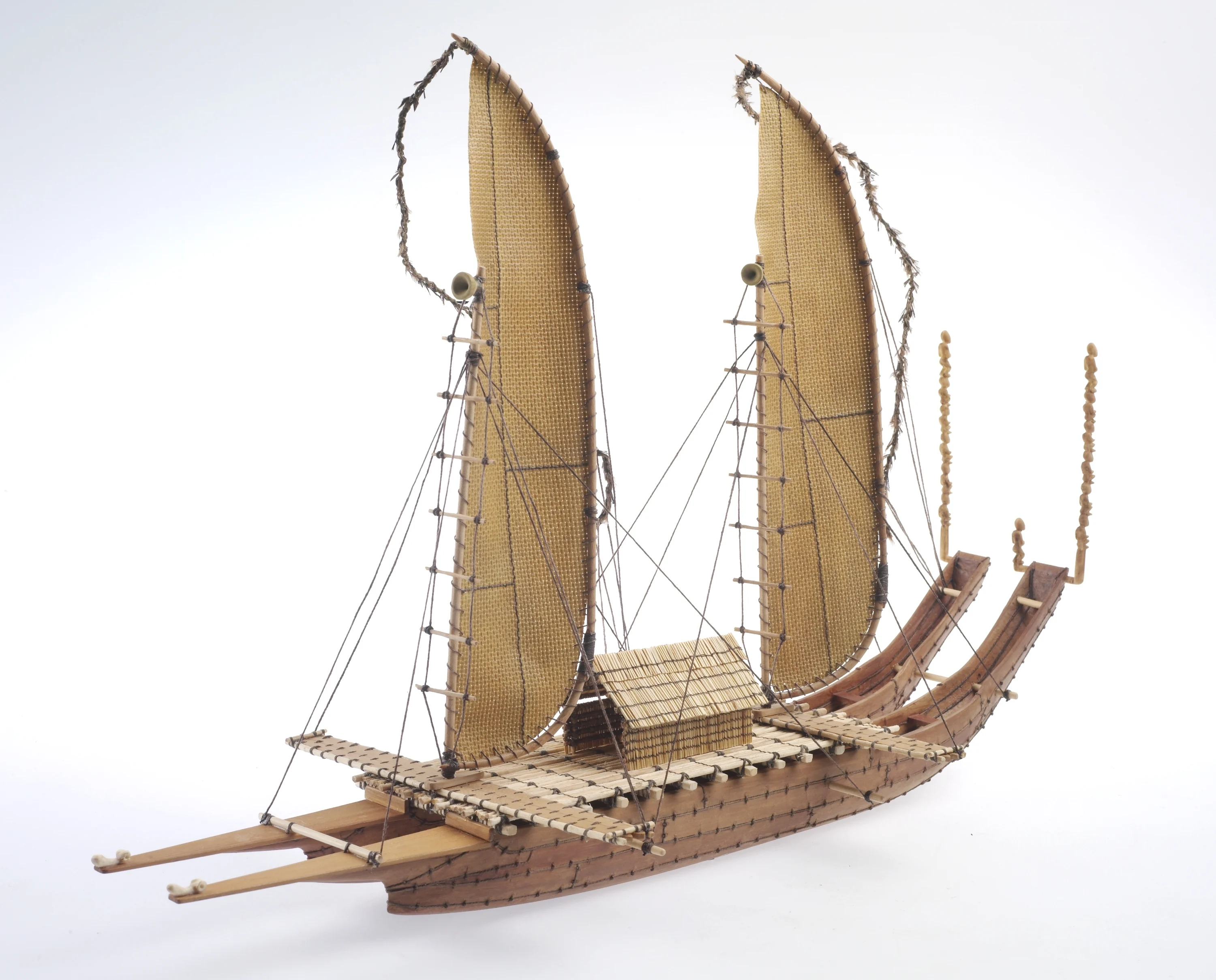 a model double hulled canoe made of wood. It has two canoe hills that curve up the back. They are joined with a connecting platform with a house like structure in the middle. It has two woven sails, and is covered in ropes and guy lines.