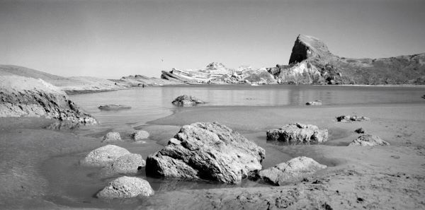 Black and white photograph over a sandy beach. The shallow water laps against the sandy shore and rough rocks, echoed by a rocky crag in the background.