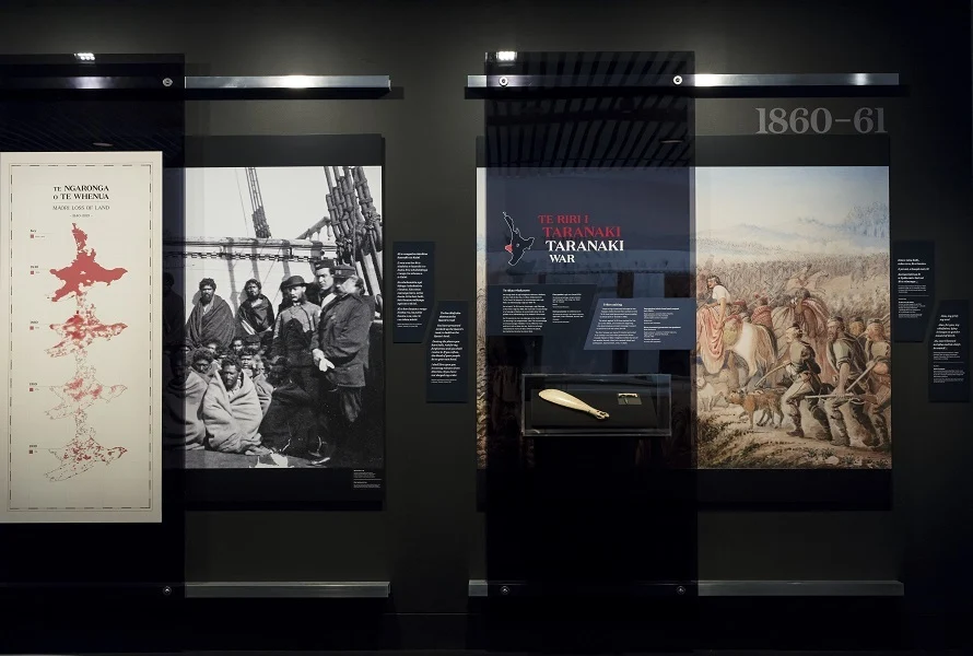 A double Museum display about the Taranaki War. There is Māori and English text on each display. On the left is a diagram showing massive loss of Māori land to British ownership. On the right, is a painting of lines of soldiers, and a glass case containing a traditional Māori whalebone club and greenstone jewellery.