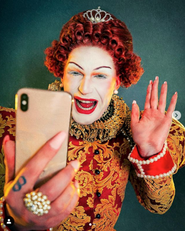 A photograph of Mary with bright red, tightly curled hair and a small crown with diamonds and pearls. Her face is pale with bright red lipstick and orange and green eye shadow. She smiles widely. She holds up a phone in one hand, to face her, and waves at it. Her dress is elaborate, with red silk and decorative orange flowers. The sleeves of the dress are puffed up at the shoulders. The is a ruffled lace collar in gold around the neckline and extending down.