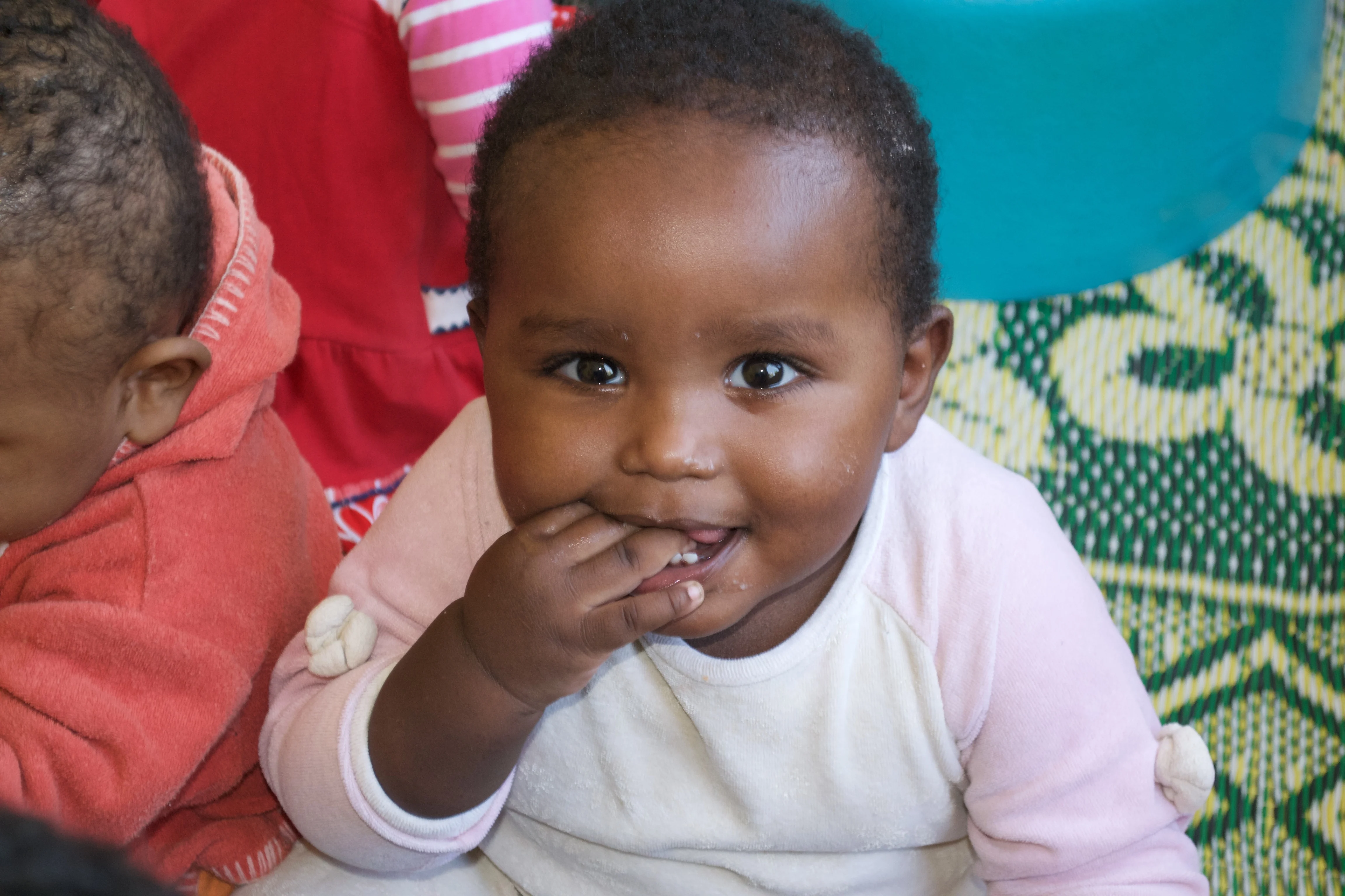 A young child on its front, smiling up at the camera with one hand in her mouth.