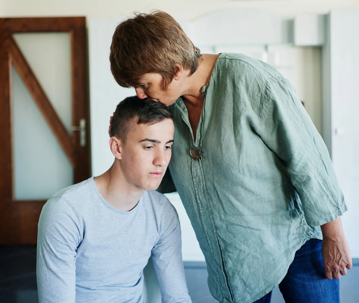 a young man sitting down next to his mother who is standing, cradling his head and kissing his forehead.