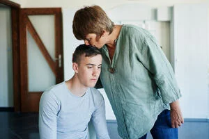 a young man sitting down next to his mother who is standing, cradling his head and kissing his forehead.