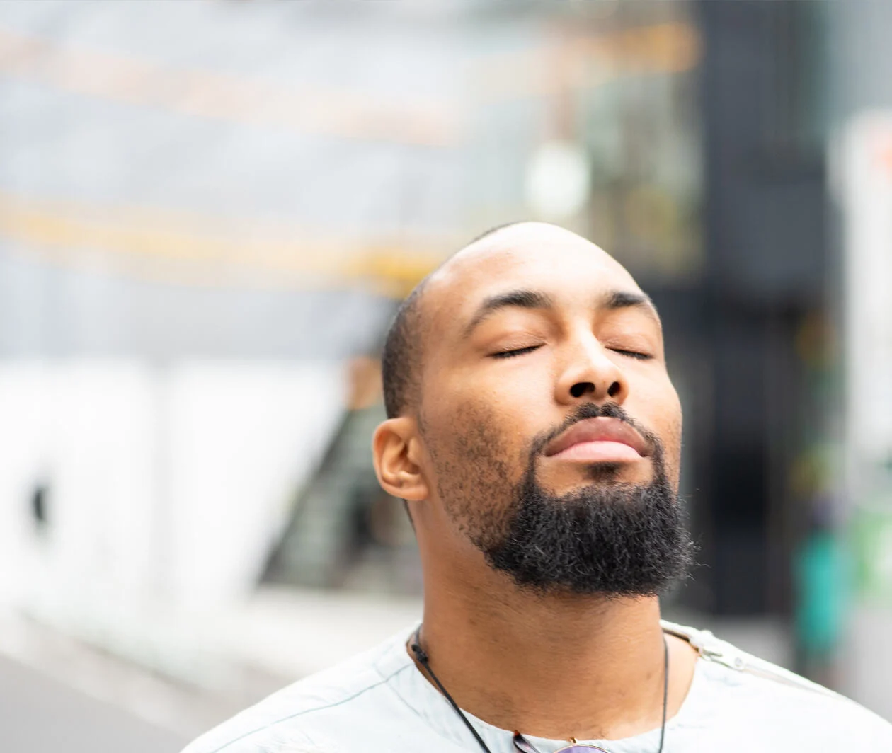 A man with beard standing on the street with his eyes closed, enjoying a moment of peace.