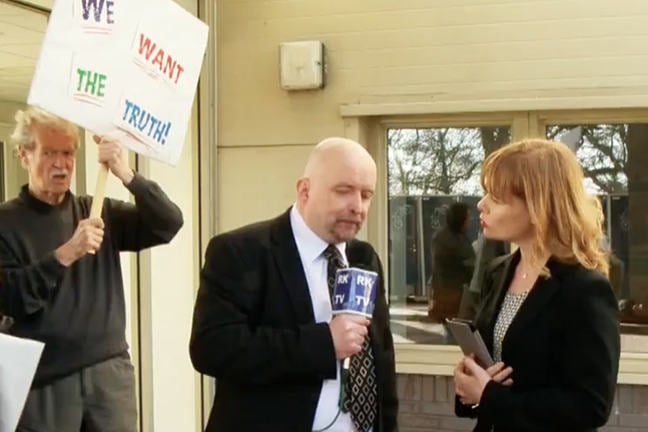 Television interviewer asking questions to a hospital representative with a protestor in the background.