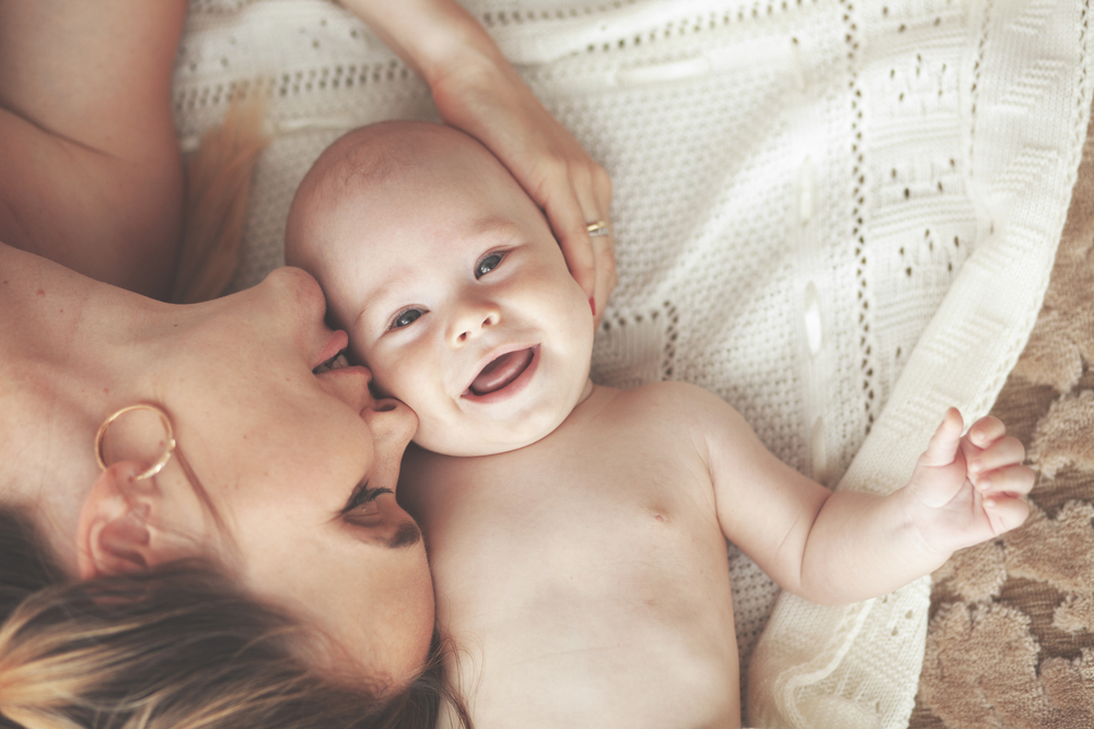 A portrait of a mother lying on a rug with her 3 month old, smiling baby.