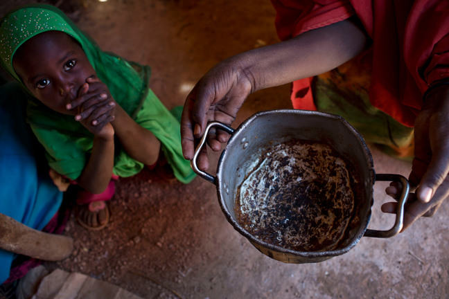 A child sits crouched on the ground with hands clasped looking up at a woman holding an empty pot of food.