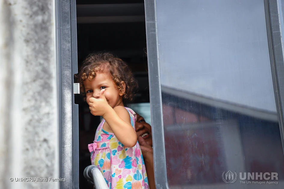 Young girl looks out of window with her thumb up smiling.