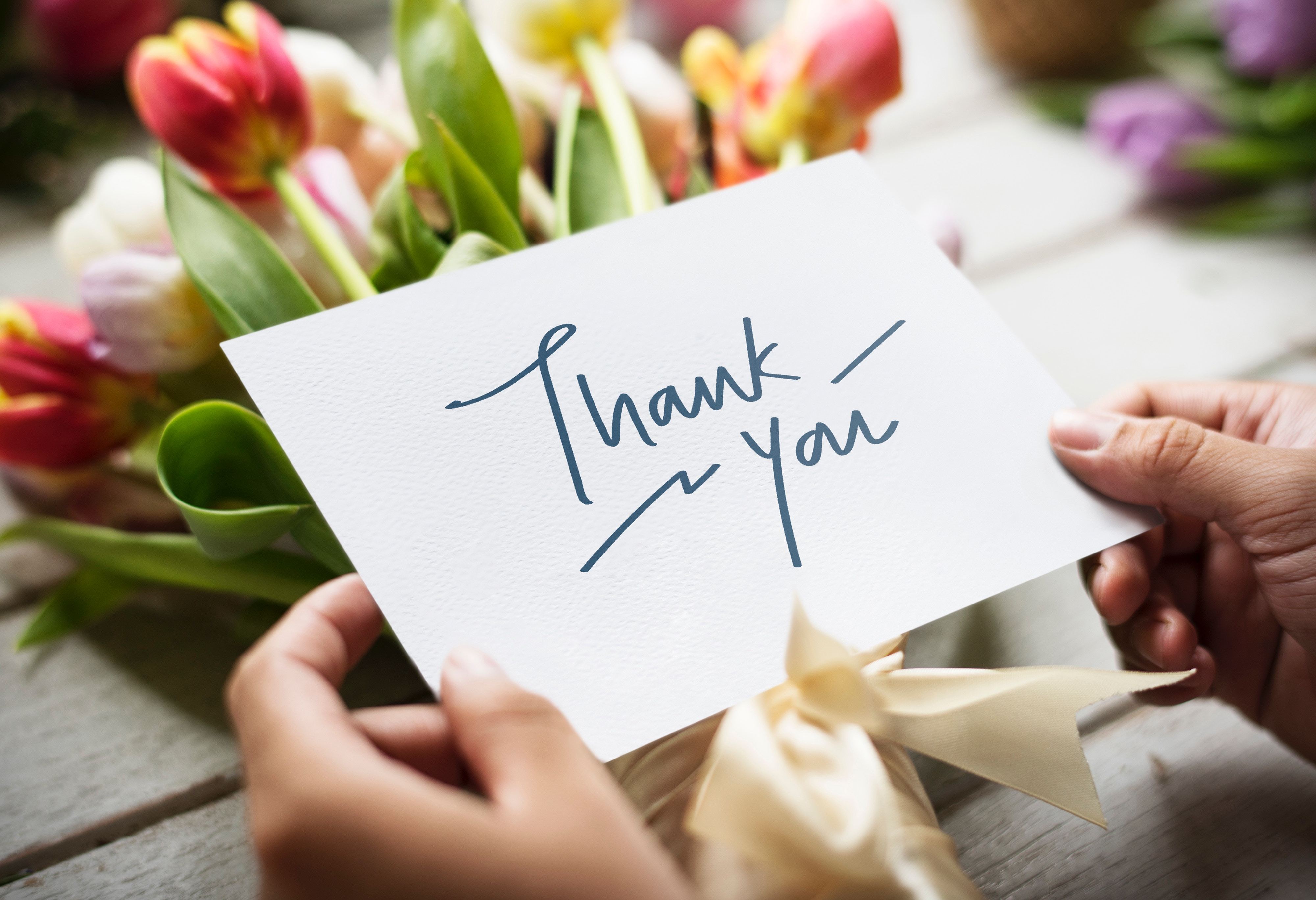 Bunch of flowers on a table with a woman standing over them reading a card that says 'Thank You'