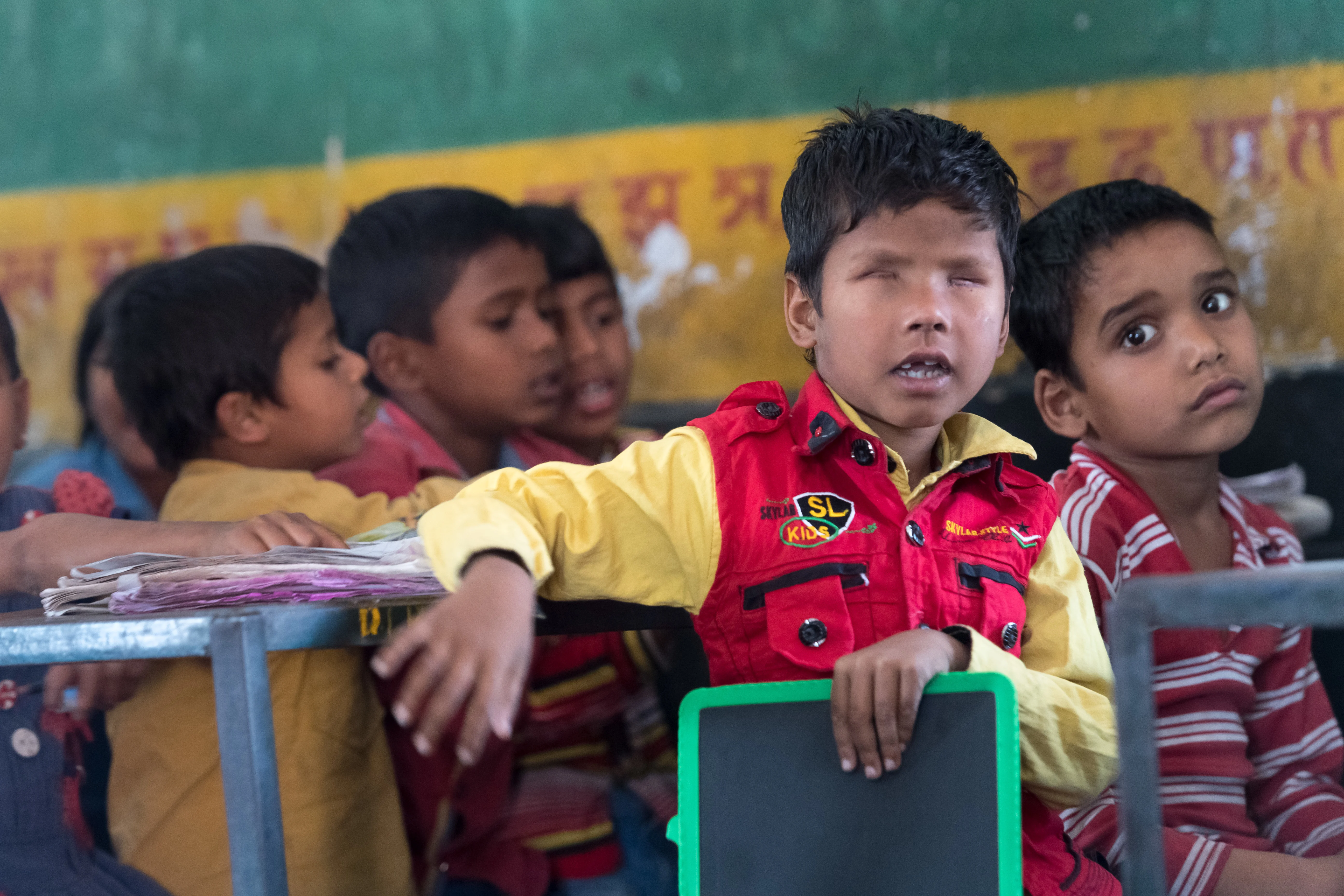 A boy born without retina is sitting with classmates at school. He is resting a hand on a chair as he turns towards the camera.