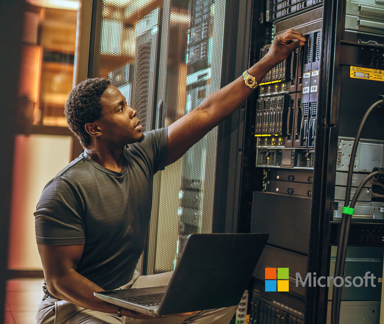 A male Azure data engineer sat in a sever room, connecting the laptop on his lap to the server tower next to him, to begin his data analytics. 