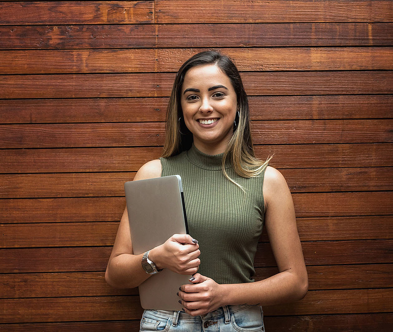 A young new graduate woman stands holding a laptop and smiling