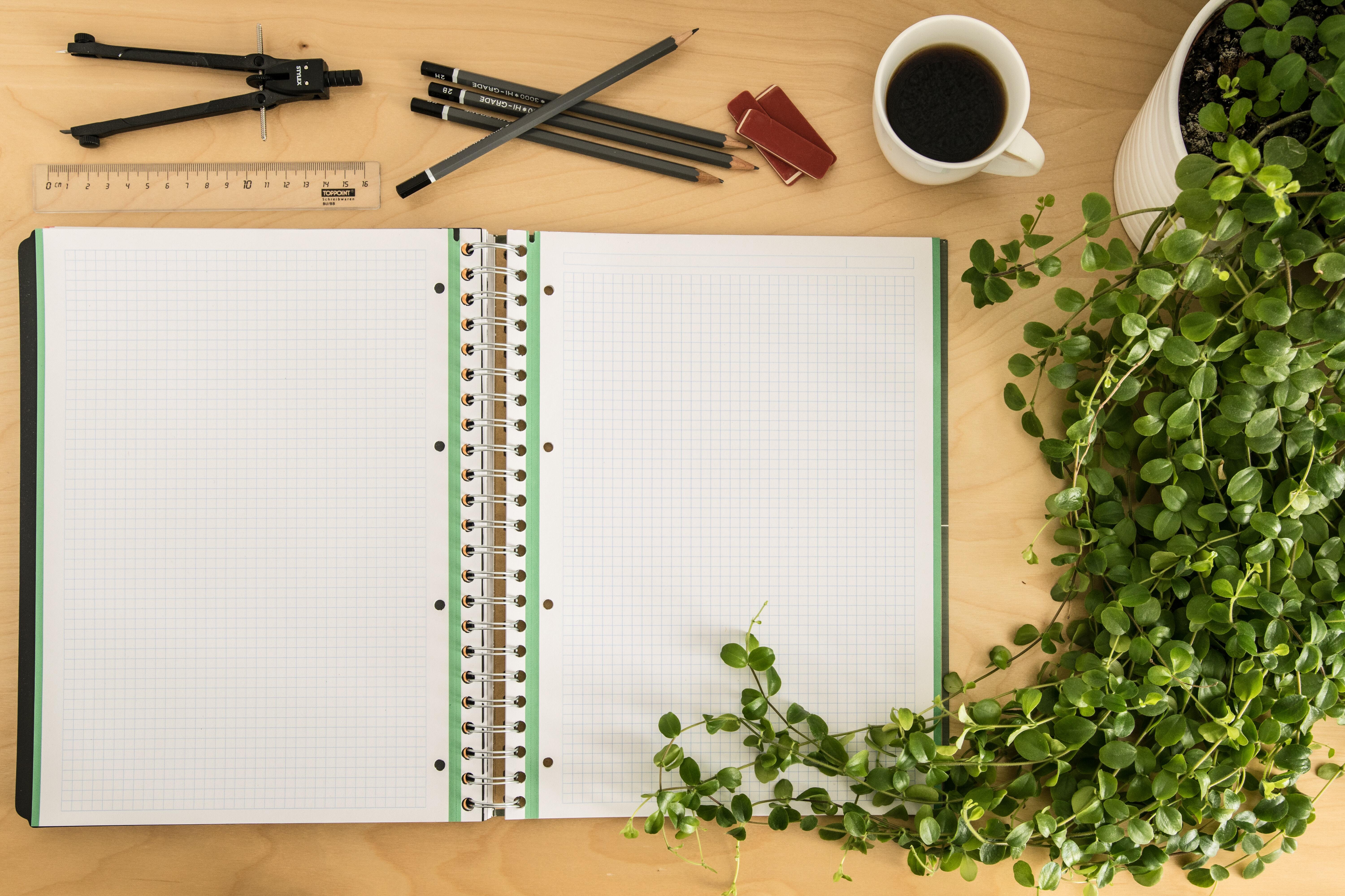 Open notebook on table with house plant, white mug of coffee, ruler pencils and mathematical compass