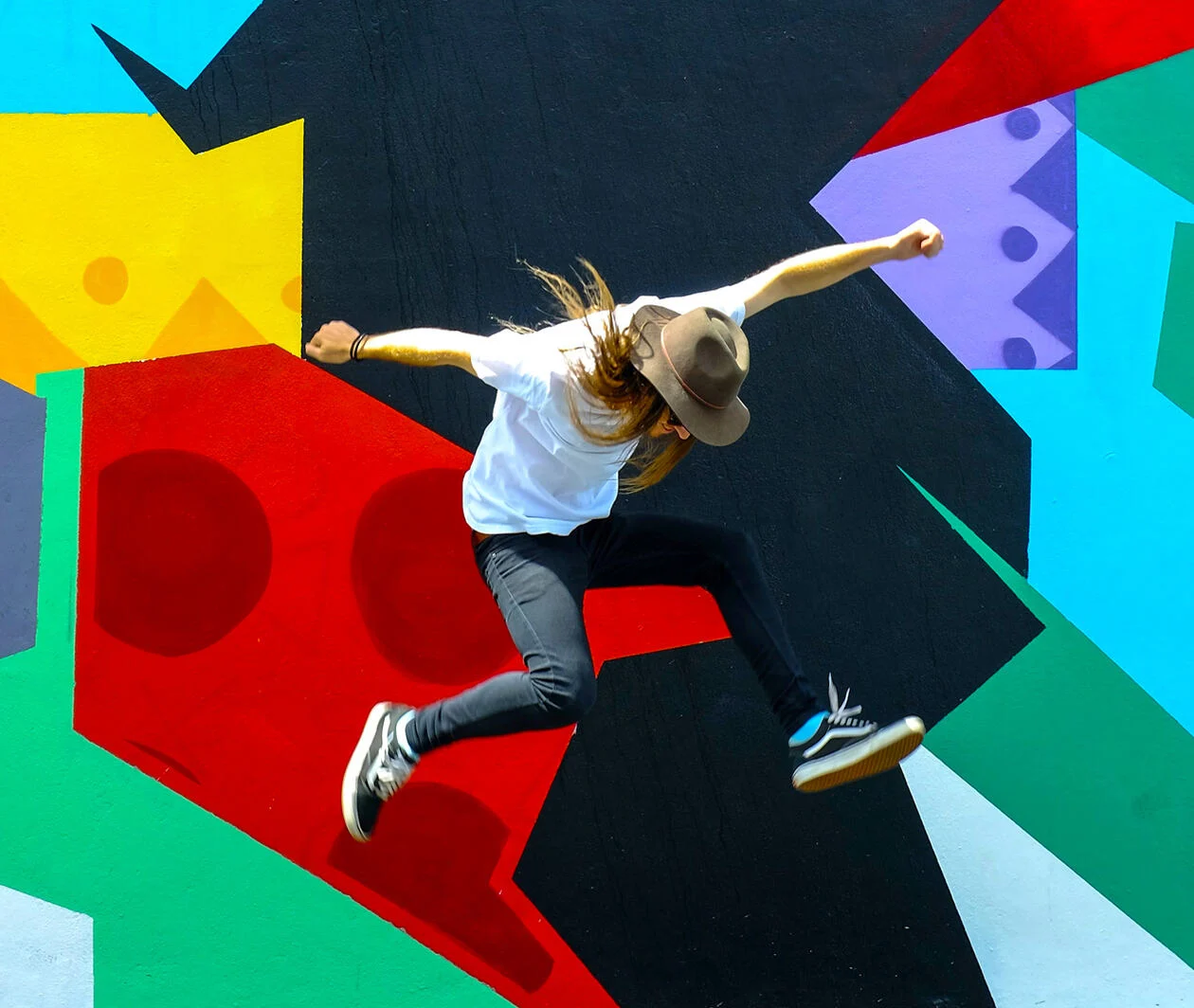 Young person with long hair jumping beside an abstract piece of graffiti