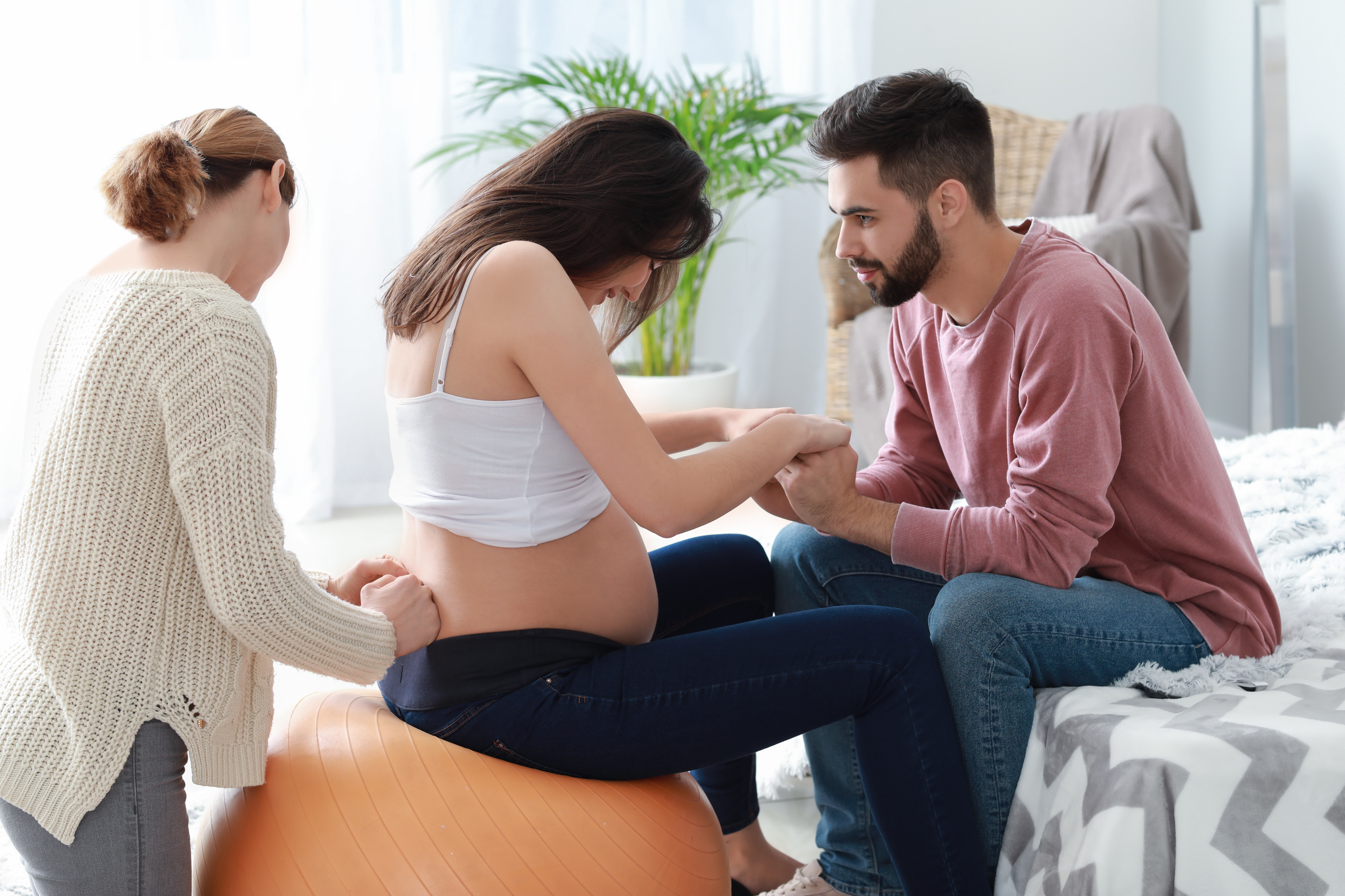 Pregnant woman on an exercise ball having her back massaged by woman and her hands held by partner