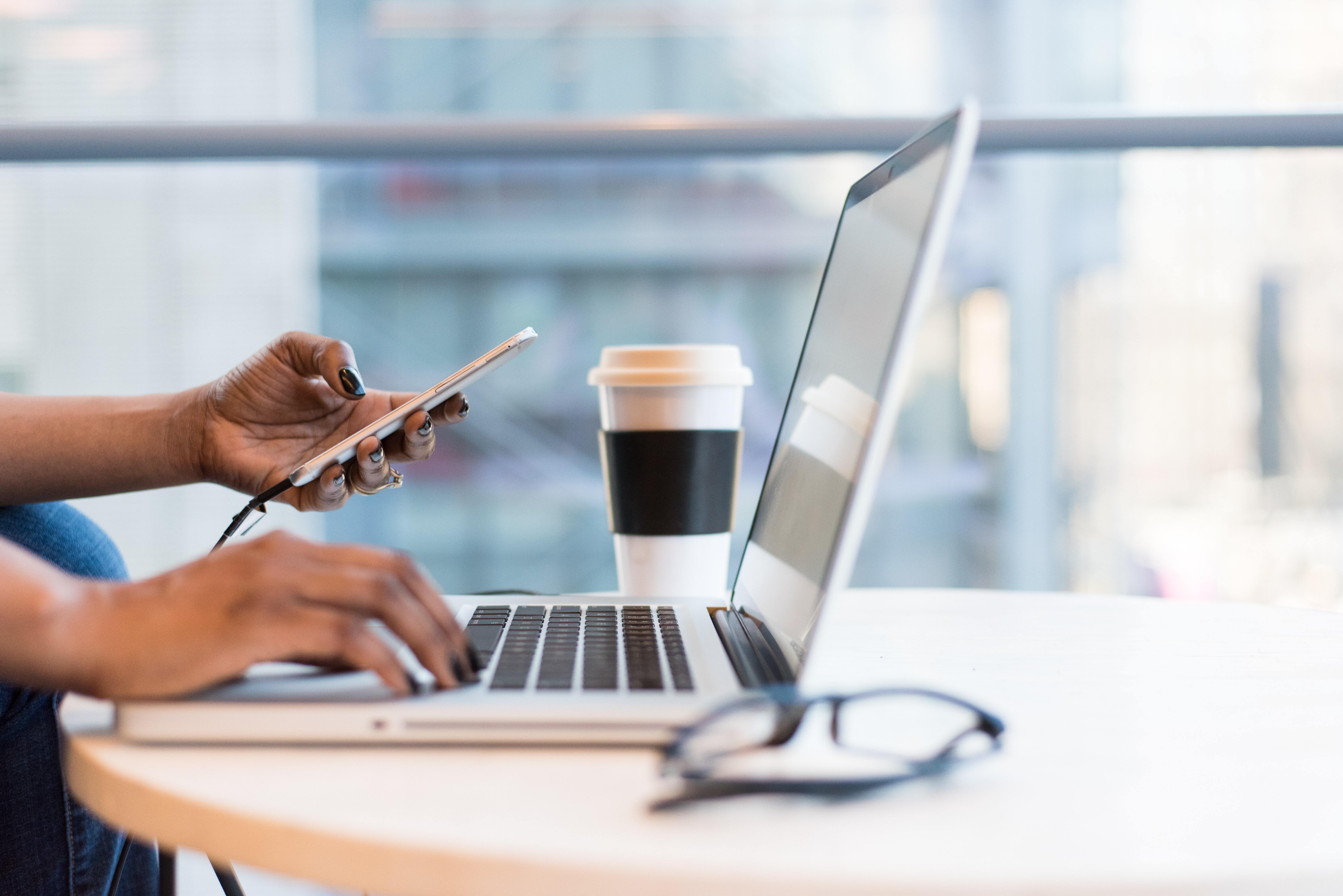 An individual working at their laptop with their phone and a hot drink.